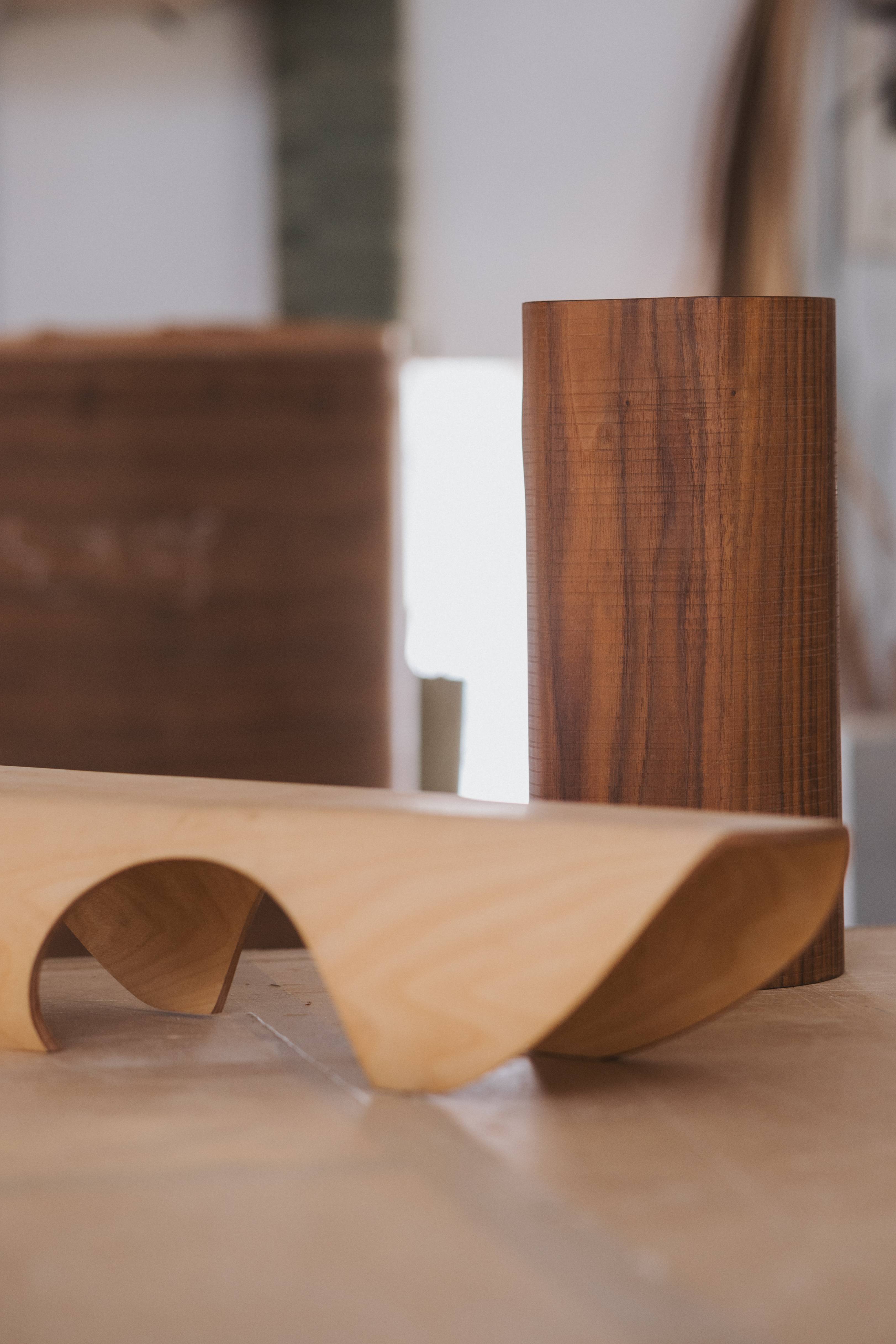 A pale bent-wood piece in a soft wave, in front of a walnut column on the workbench.